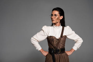 pensive steampunk woman in glasses standing with hands on hips isolated on grey