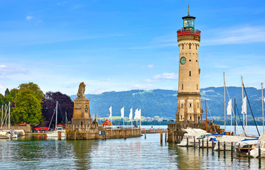 Lindau, Germany. Old lighthouse with clock in bay. Antique bavarian town at Lake Constance (Bodensee). Monument with statue of lion at entrance to port, yachts by piers. Summer landscape blue sky.