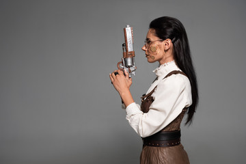 Wall Mural - side view of attractive steampunk woman holding vintage pistol isolated on grey