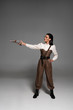 © LIGHTFIELD STUDIOS - full length view of attractive steampunk woman standing with hand on hip and aiming with pistol on grey