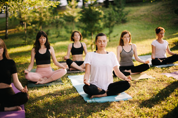 Group of mixed age women is practicing yoga and meditating morning in city park while sunrise. Group of people is sitting outdoors in lotus pose on grass with eyes closed