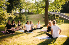 Group Of Smiling Women Prepare To Meditation In Park On Summer Sunny Morning Under Guidance Of Trainer. Group Of Girl Are Sitting In Lotus Pose On Yoga Mats Are Relaxing Talking Before Yoga Training