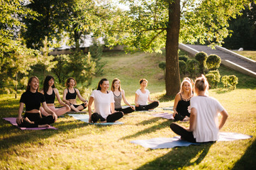 Group of smiling women prepare to meditation in park on summer sunny morning under guidance of trainer. Group of girl are sitting in lotus pose on yoga mats are relaxing talking before yoga training