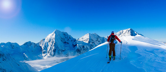 Fototapete - Mountaineer backcountry ski walking up along a snowy ridge with skis in the backpack. In background blue sky and shiny sun and Zebru, Ortler in South Tirol, Italy.  Adventure winter extreme sport.