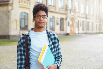 Wall Mural - Smiling African American student with glasses and with books near college. Portrait of a happy black young man standing on a university background.