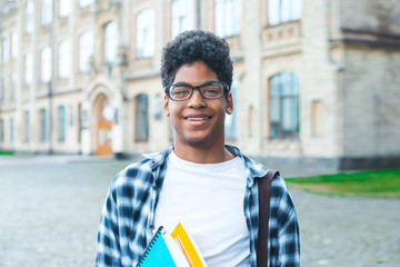 Wall Mural - Smiling African American student with glasses and with books near college. Portrait of a happy black young man standing on a university background.