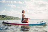 Middle age Caucasian woman practising yoga on paddle sup surfboard at sunset. Female stretching doing workout on lake water. Modern individual hipster outdoor summer sport activity.