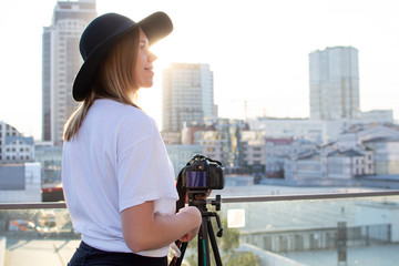 Canvas Print - girl photographer with a camera and a tripod on a background of the city, she photographs at sunset, a woman shoots a video