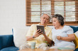 © tirachard - Asian elderly couple using tablet and drinking coffee in living room at home, couple enjoy love moment while lying on sofa when relaxed at home. Enjoying time lifestyle senior family at home concept.