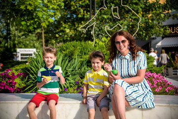 Wall Mural - Tasty summer obsession concept. Happy young family boys with mother wearing colorful clothes, eating mini melts ice cream in heat cap over summer city park green nature plants background