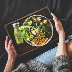Wall Mural - Healthy dinner, lunch in bed. Vegan superbowl or Buddha bowl with hummus, vegetable, fresh salad, beans, couscous and avocado, green smoothie on tray and woman in jeans eating, top view, square crop