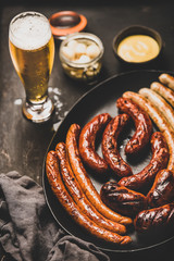 Wall Mural - Octoberfest dinner table concept with grilled veal and pork sausages, sauce in jar, pickled vaggies and lager beer in glass over dark background, selective focus, close-up