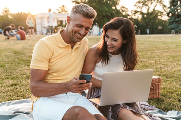 Wall Mural - Portrait of smiling middle-aged couple holding cellphone while using laptop computer during picnic in summer park