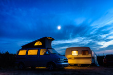 A Camper Van With Its Pop Top Roof Open Parked Next To A Vintage Caravan In A Campsite At Nightfall, Both Illuminated From Inside And The Moon Glowing Above In A Stormy Sky.