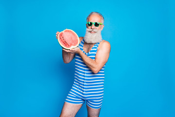 Wall Mural - Portrait of retired pensioner in eyeglasses eyewear holding tropical exotic fruit wearing striped bathing suit isolated over blue background