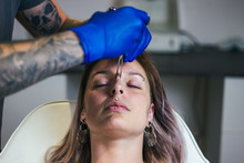 Young Woman Getting Pierced Between Her Eyes. Man Showing A Process Of Piercing With Steril Neadle And Latex Gloves. Body Piercing Procedure