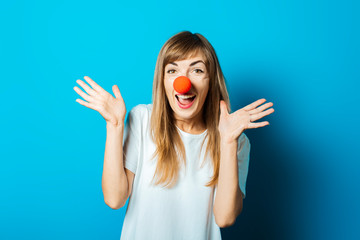 Beautiful young woman in a white T-shirt and a red clown nose smiles and makes hand gestures on a blue background. Concept party, costume, red nose day