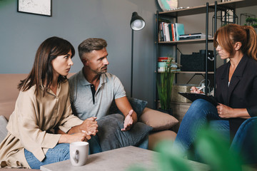 Photo of serious brooding couple having conversation with psychologist on therapy session in room