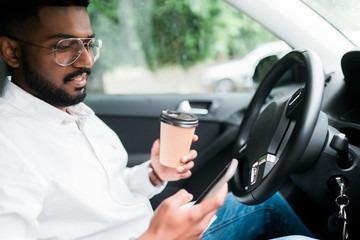 Indian man driver with a smartphone in hand and paper cup of hot coffee. The concept of inattention at the wheel, rest, coffee break to cheer.