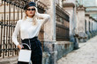 © Victoria Fox - Outdoor street fashion portrait of elegant lady wearing stylish autumn outfit: black leather beret, sunglasses, white turtleneck, checkered high waist trousers, small bag, posing in city. Copy space