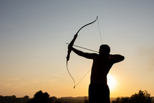 Silhouette Of A Man With An Ancient Weapon Bow And Arrow On A Background Of Sky And Sunset