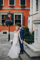 Wall Mural - Stylish bride and groom kissing sensually in sunny european city street. Gorgeous wedding couple of newlyweds embracing at old buildings. Romantic moment