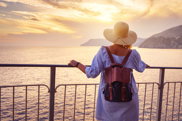 Woman in casual dress and hat with backpack looking to sea. Travel Cinque Terre in Italy in summer sunset