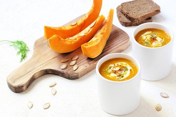 Roasted pumpkin and carrot soup with cream and pumpkin seeds on two white mug on white background with black bread slices.Vegetarian eating.Diet and healthy food concept.Space for copy and text