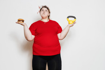 Wall Mural - Fat and funny girl dressed in the sportswear and with a bandage on her head is standing with a burger in the one hand and the weight in the other against a white wall