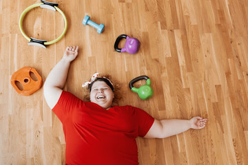 Wall Mural - Laughing fat girl dressed in the sportswear and with a bandage on her head is laying on the floor next to the sports equipment