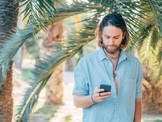 Young bearded hipster man wearing blue shirt texting on phone in tropical jungle