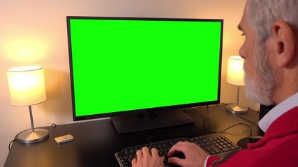 An elderly man works on a computer with a green screen in an apartment - closeup - types on keyboard