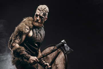 Wall Mural - Medieval warrior berserk Viking with tattoo on skin, red beard and braids in hair with axe and shield attacks enemy. Concept historical photo