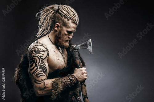 Fotografía Medieval warrior berserk Viking with tattoo on skin, red beard and braids in hair with axe and shield attacks enemy