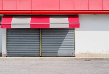 Shop Retail With Metal Shutters Closed And A Red White Awning. Sidewalk And Asphalt Road In Front. Urban Background For Copy Space