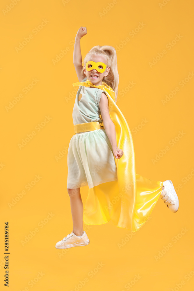 Jumping little girl dressed as superhero on color background