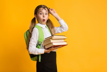 Shocked Primary School Girl Holding Stack Of Books, Yellow Background