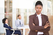 © Mangostar - Stressed African American female employee standing with arms crossed and looking away. Her male and female colleagues sitting in background. Successful business woman concept