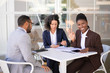 © Mangostar - Happy African American business woman waving hello, while her colleagues discussing work issues. Multiethnic business man and women sitting in outdoor. Work break concept