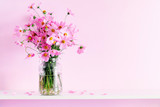 Fototapeta Kosmos - Fresh summer bouquet of pink cosmos flowers in glass vase on white wood shelf on pink wall background. Floral home decor.