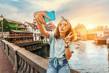 Wall Mural - Happy and cheerful Asian girl traveler takes a selfie on the Saint Martin bridge in the Petit France area in Strasbourg. Colorful and dramatic sunset at the background