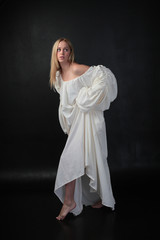 Wall Mural - full length portrait of blonde girl wearing long white flowing robe. standing pose against a black studio background.