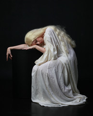 Ghostly  portrait of a woman with long blonde hair wearing a white robe. posing against a black studio background. 