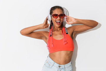 Wall Mural - Positive optimistic young african stylish woman in sunglasses posing isolated over white wall background listening music with headphones.