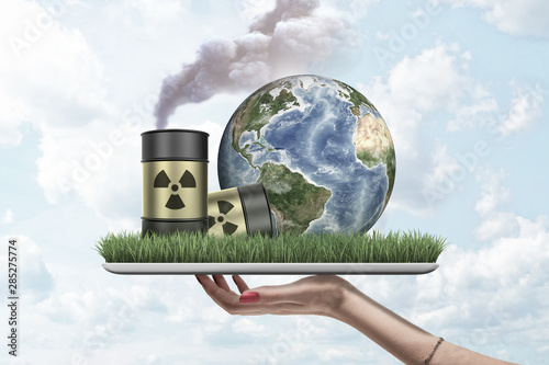 Cuadros en Lienzo Woman's hand holding digital tablet with green grass, planet Earth and two radioactive waste barrels placed on screen, against cloudy sky