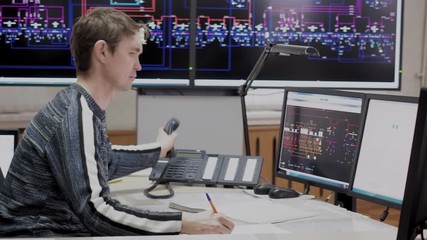 Office with digital screens with schemes tracking electricity objects. Man system administrator working on computer control room of electric power station. He makes paper work and answer phone calls.