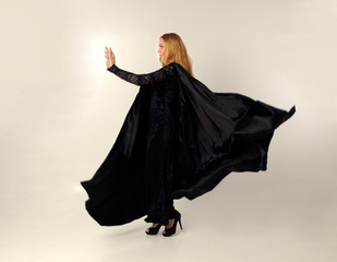 Wall Mural - full length portrait of blonde woman wearing long black flowing clock and lace gown, standing pose against a creamy studio background.