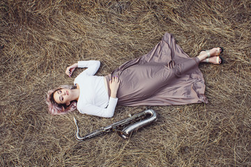 Wall Mural - body of a beautiful young woman in long dress lying on the dry grass next to the saxophone, blonde girl rests on the hay with a musical instrument, top view