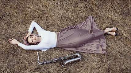 Wall Mural - body of a beautiful young woman in long dress lying on the dry grass next to the saxophone, blonde girl rests on the hay with a musical instrument, top view