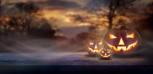 Mist rolling in over the spooky glow of Jack O' Lanterns on the right hand side of a wooden bench in a forest at dusk.